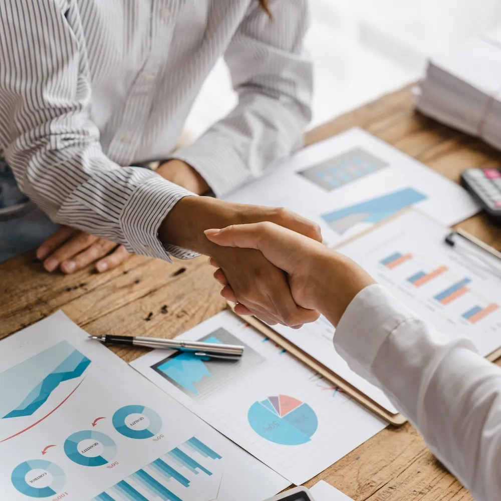 two people shaking hands over sheets of financial graphs and charts
