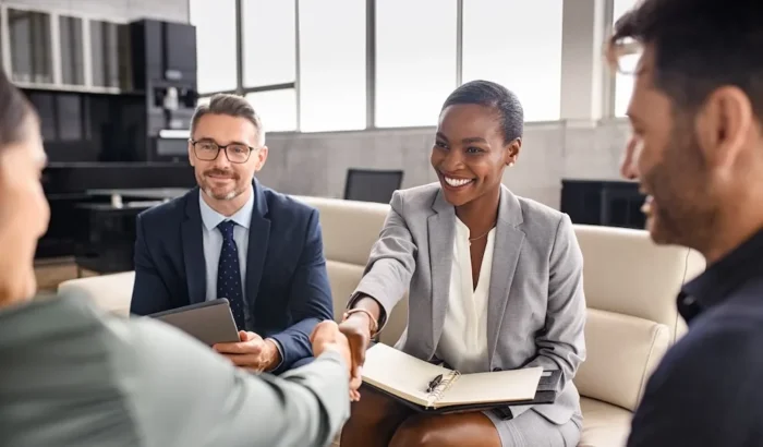 The focus is on a Black businesswoman, seated on a couch with 2 businessmen, shaking hands with a person across from her in a high end office setting