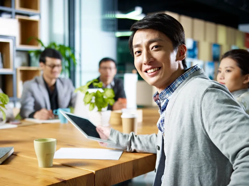 seated Asian man smiling and turning away from the work table with other business people
