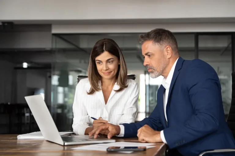 A businesswoman and businessman are seated next to each other both looking at a laptop