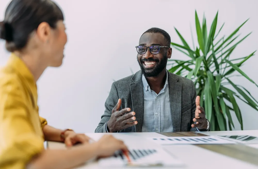 a Black man is talking to an Asian woman in an office setting with sheets of colored financial charts on the desk