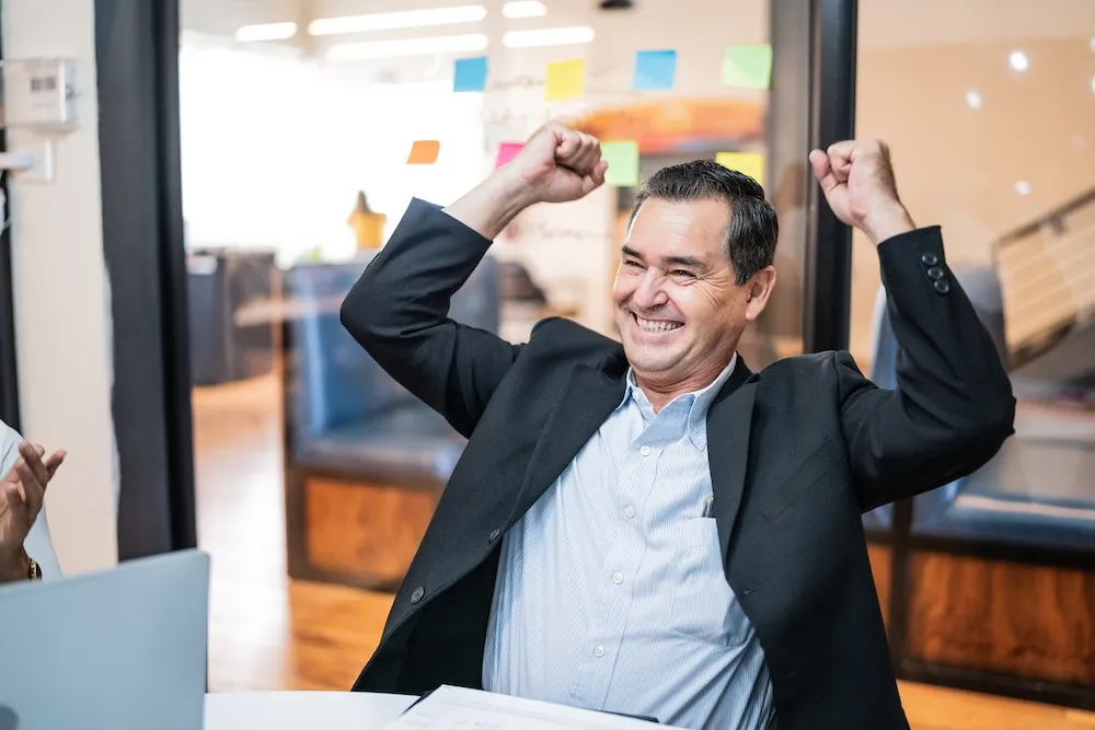 A happy White business exec has his arms and fists raised in a celebratory movement