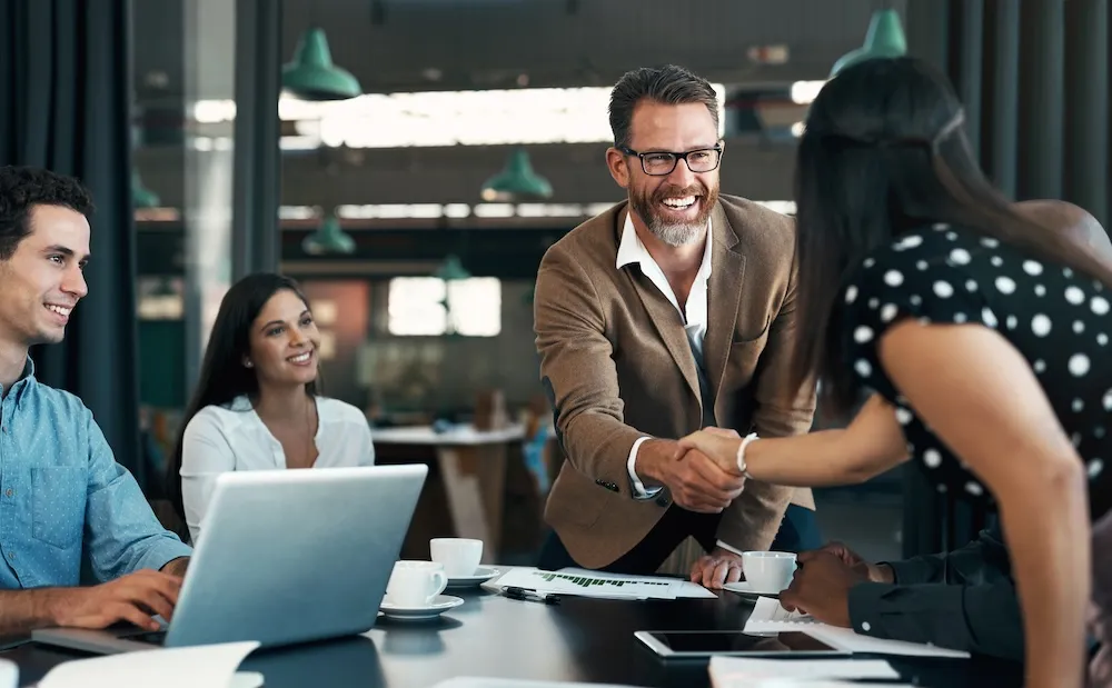 A White businessperson is standing up from the table cordially shaking hands with a woman show is with others seated at the table
